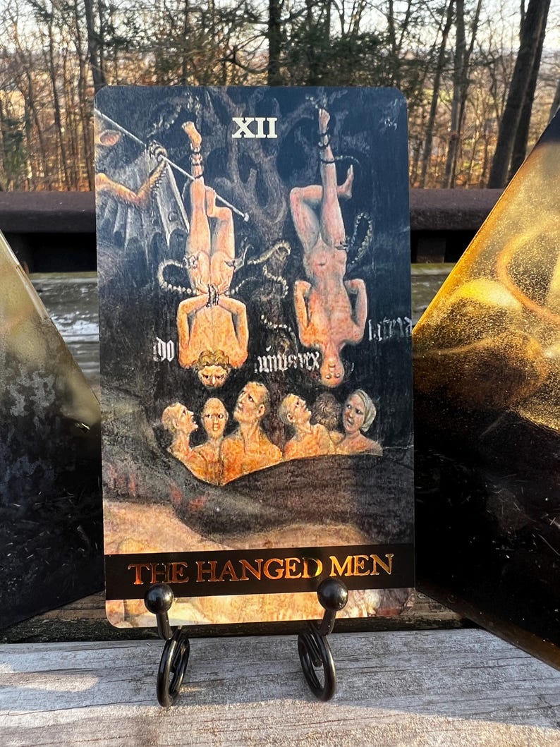 May include: Tarot card featuring an illustration of two figures hanging upside down, with a group of people below. The card is labeled "XII" and "THE HANGED MEN" in gold lettering. The card is displayed on a black metal stand.