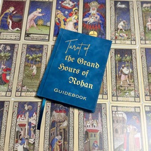 May include: A teal blue hardcover book with gold foil lettering that reads Tarot of the Grand Hours of Rohan GUIDEBOOK. The book features a matching teal ribbon bookmark and is positioned over a collection of rectangular tarot cards.