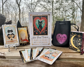 Op de afbeelding: Een Cat Folk Tarot-deck met 78 kaarten en een 161 pagina's tellend handboek. Het deck bevat illustraties van katten in verschillende poses en omgevingen. De kaarten zijn gerangschikt op een houten tafel met een zwarte tas en een enkele kaart die een katten silhouet tegen een halve maan laat zien.