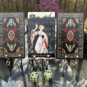 May include: Three tarot cards with a gothic design and a central card featuring a woman. The cards are black with red pentagrams and bat illustrations. The central card reads "Inspiration Fuels My Journey". Two green dice are in the foreground.