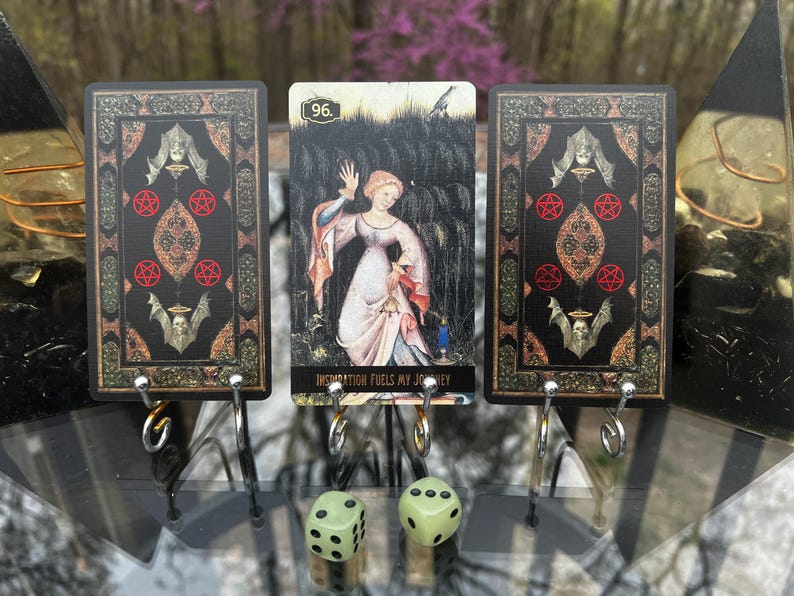 May include: Three tarot cards with a gothic design and a central card featuring a woman. The cards are black with red pentagrams and bat illustrations. The central card reads "Inspiration Fuels My Journey". Two green dice are in the foreground.