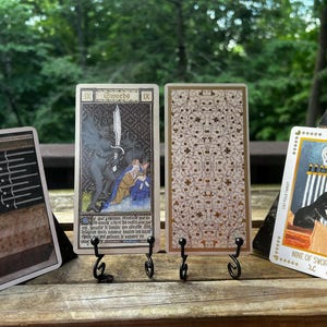 May include: Four tarot cards displayed on small black metal stands. The left card shows a person in bed with nine swords above. The second card depicts a dark figure with a sword. The third card has a floral pattern. The right card shows a cat with nine swords and the text Nine of Swords.