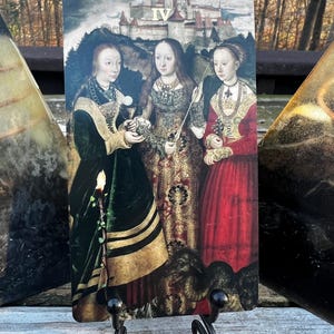 May include: A tarot card featuring a Renaissance-style painting of three women in ornate gowns, with a castle in the background. The card is held by a black metal stand. The Roman numeral IV is visible at the top.