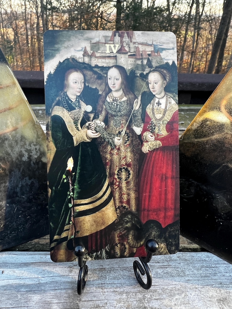 May include: A tarot card featuring a Renaissance-style painting of three women in ornate gowns, with a castle in the background. The card is held by a black metal stand. The Roman numeral IV is visible at the top.