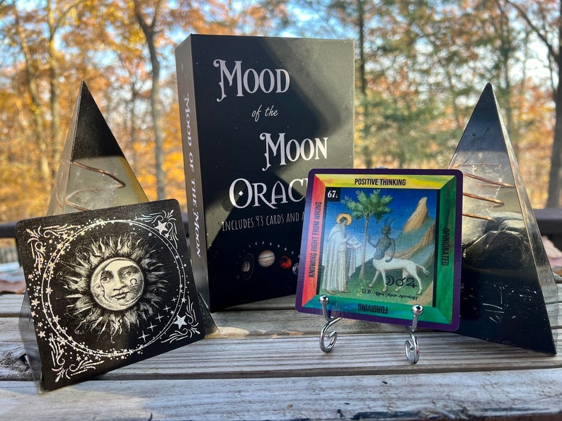 Może przedstawiać: Czarne pudełko z napisem "Mood of the Moon Oracle" i tęczowa karta z napisem "Positive Thinking" i numerem "67". Karta stoi na srebrnym stojaku. Po obu stronach pudełka znajdują się dwie czarne piramidy z kryształu.