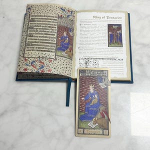 May include: An open book with medieval-style illuminated text and illustrations on the left page and the title King of Pentacles on the right page. A rectangular tarot card featuring a king in blue robes sitting with a bull is placed below.