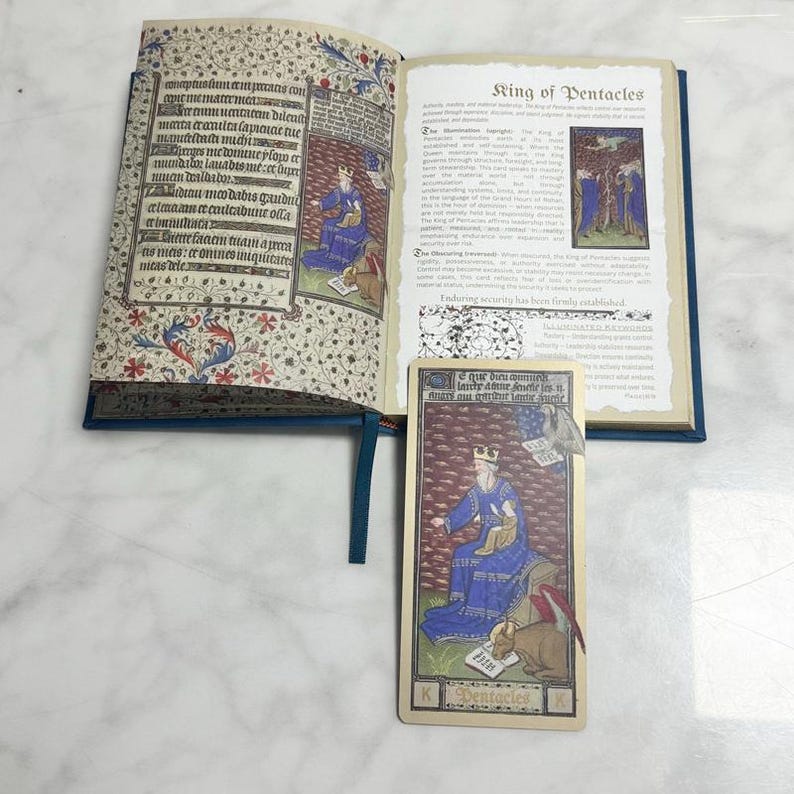 May include: An open book with medieval-style illuminated text and illustrations on the left page and the title King of Pentacles on the right page. A rectangular tarot card featuring a king in blue robes sitting with a bull is placed below.