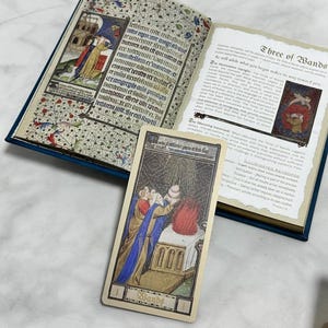 May include: An open book with illuminated manuscript-style illustrations and text, accompanied by a single tarot card titled Three of Wands. The card features a figure in blue and gold robes standing before a stone altar with a red flame.