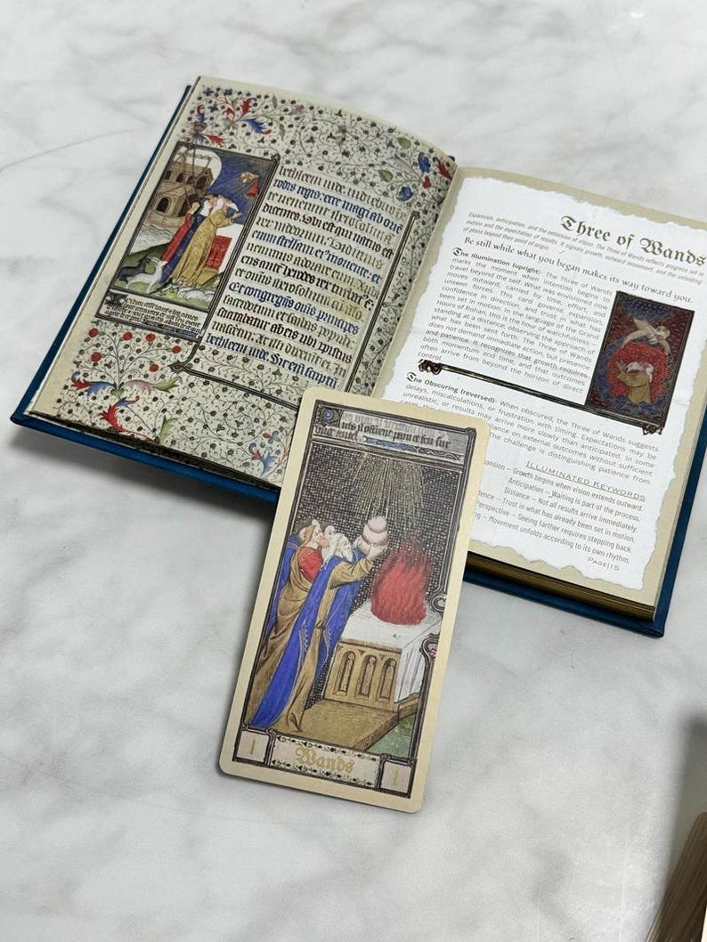 May include: An open book with illuminated manuscript-style illustrations and text, accompanied by a single tarot card titled Three of Wands. The card features a figure in blue and gold robes standing before a stone altar with a red flame.
