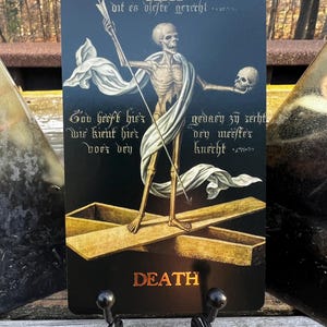 May include: A tarot card featuring a skeletal figure holding a staff and a skull, standing on a golden cross. The card has the word "DEATH" in gold lettering at the bottom. The background is dark blue with script.