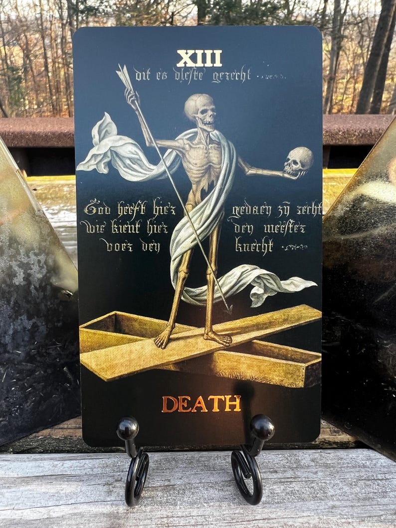 May include: A tarot card featuring a skeletal figure holding a staff and a skull, standing on a golden cross. The card has the word "DEATH" in gold lettering at the bottom. The background is dark blue with script.