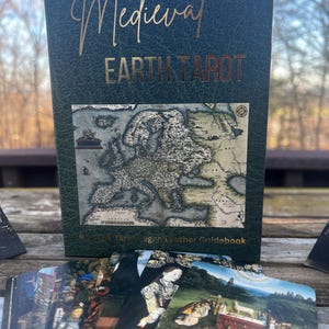 May include: A Medieval Earth Tarot deck with a dark green cover and gold lettering. The cover features a map of Europe and the deck is accompanied by a guidebook. The cards are visible in the foreground, displaying various illustrations.