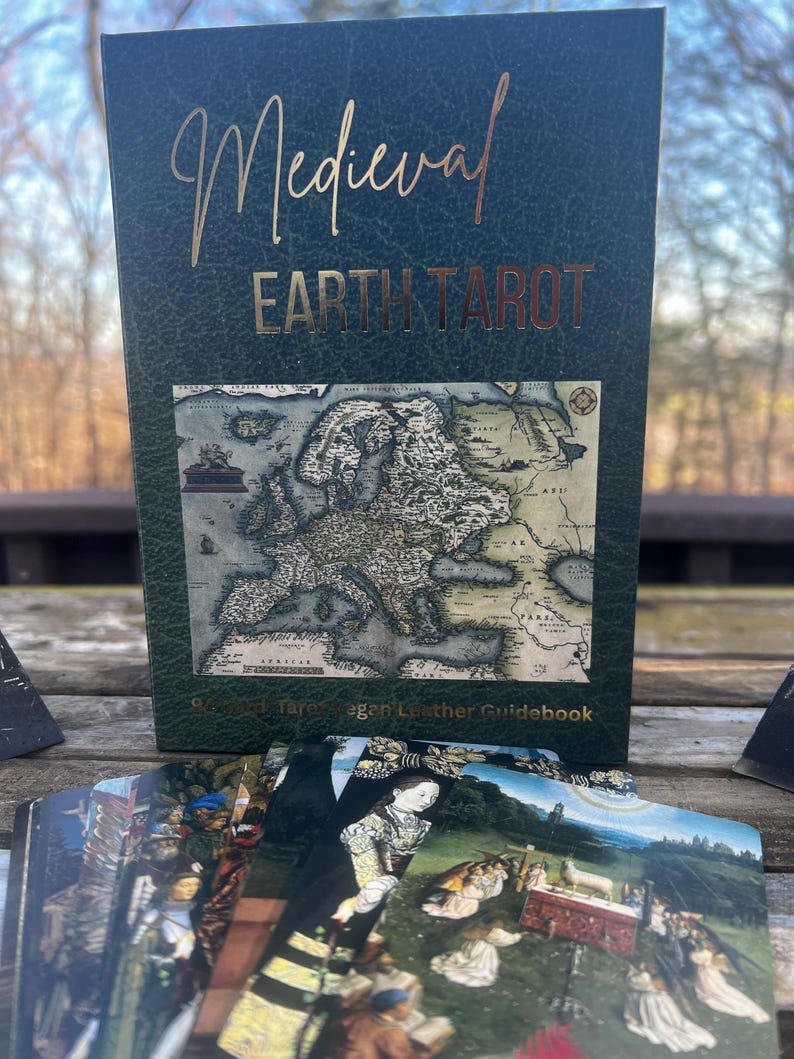 May include: A Medieval Earth Tarot deck with a dark green cover and gold lettering. The cover features a map of Europe and the deck is accompanied by a guidebook. The cards are visible in the foreground, displaying various illustrations.