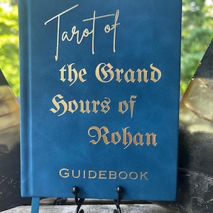 May include: A blue hardcover guidebook with gold foil lettering on the front cover that reads Tarot of the Grand Hours of Rohan. The book features a smooth matte finish and a thin blue ribbon bookmark extending from the bottom edge of the spine.