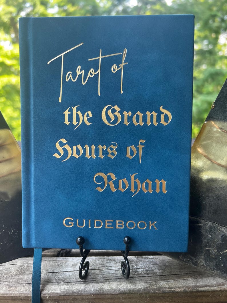 May include: A blue hardcover guidebook with gold foil lettering on the front cover that reads Tarot of the Grand Hours of Rohan. The book features a smooth matte finish and a thin blue ribbon bookmark extending from the bottom edge of the spine.