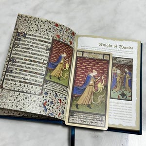 May include: An open book with illuminated manuscript-style pages featuring intricate floral borders and Latin text. A rectangular tarot card titled Knight of Wands, depicting a figure in a blue cloak and tan robe with a dragon, rests on the right page.