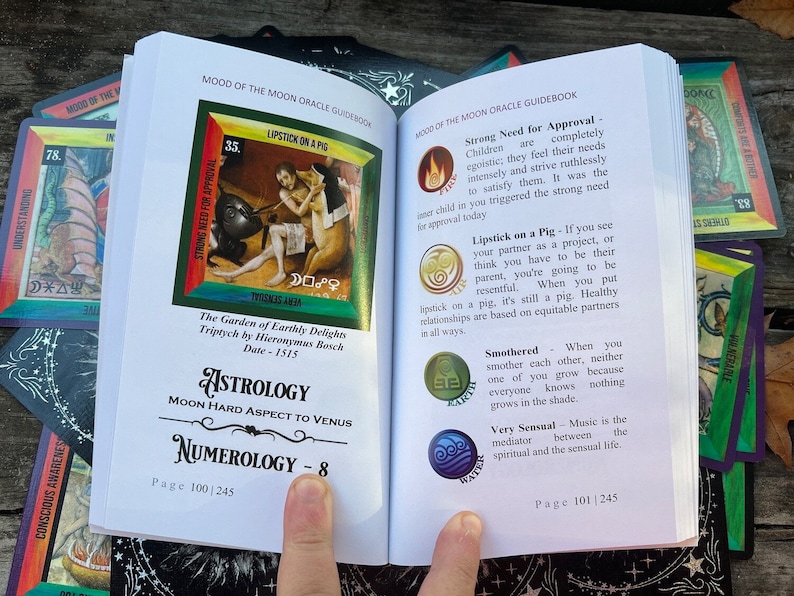 Może przedstawiać: Otwarta książka z kolorową ilustracją kobiety nakładającej szminkę na świnię. Książka nosi tytuł "Mood of the Moon Oracle Guidebook" i zawiera tekst o astrologii i numerologii. Książka jest otwarta na stronach 100 i 101.