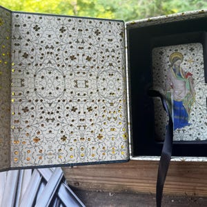 May include: A rectangular box with a hinged lid, featuring an intricate cream and gold floral pattern on the interior. Inside, a deck of cards with a religious figure illustration rests in a dark blue velvet-lined compartment with a black ribbon.