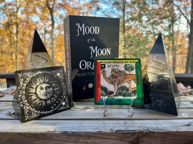 Może przedstawiać: Czarna książka zatytułowana "Mood of the Moon Oracle" z czarno-białą kartą przedstawiającą słońce i księżyc. Zielona i ż&oacute;łta karta z ilustracją lwa i tekstem "What we want conflicts with what we need" jest wyświetlana na srebrnym stojaku. Po obu stronach książki znajdują się dwie czarne i przezroczyste piramidy z kryształu.