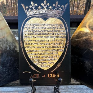 May include: A black Ace of Cups tarot card with gold accents, featuring a heart-shaped design and ornate text. The card is displayed on a black metal stand, with the words "ACE of CUPS" at the bottom.