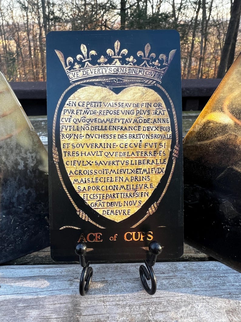 May include: A black Ace of Cups tarot card with gold accents, featuring a heart-shaped design and ornate text. The card is displayed on a black metal stand, with the words "ACE of CUPS" at the bottom.