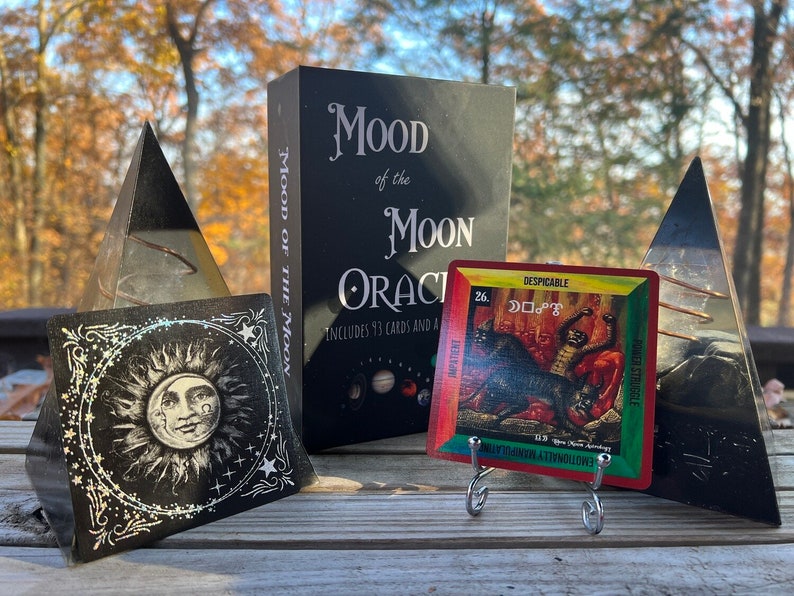 Może przedstawiać: Czarne pudełko z tekstem "Mood of the Moon Oracle" i "Includes 93 cards and a guidebook" na przodzie. Czarno-biała karta z wzorem słońca i księżyca znajduje się po lewej stronie. Kolorowa karta z czarnym kotem i tekstem "Despicable" i "26." znajduje się po prawej stronie. Karty leżą na drewnianej powierzchni.