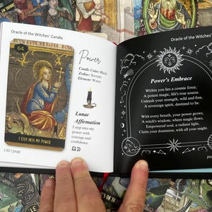 May include: An open book displaying tarot cards. One page features a card with a woman in blue and gold, the text "I STEP INTO MY POWER", and the number 64. The opposite page has the text "Power's Embrace" with a sun and moon design.