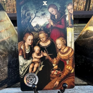 May include: A tarot card featuring a Renaissance-style painting of several figures, including a baby, women in ornate gowns, and a woman praying. The card is held by a black metal stand. The card has the Roman numeral X and a pentacle symbol.