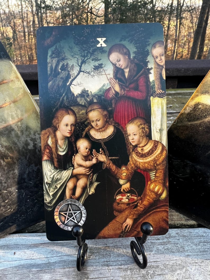 May include: A tarot card featuring a Renaissance-style painting of several figures, including a baby, women in ornate gowns, and a woman praying. The card is held by a black metal stand. The card has the Roman numeral X and a pentacle symbol.