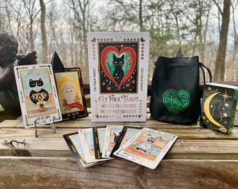 Könnte beinhalten: Ein schwarzes und grünes Tarot-Deck mit Katzenmotiv. Die Schachtel zeigt eine schwarze Katze mit einem herzförmigen Design. Das Deck enthält 78 Karten und ein 161-seitiges Handbuch. Die Karten liegen auf einer Holzoberfläche mit einer schwarzen Tasche und einer einzelnen Karte.