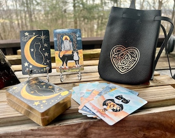 May include: A set of tarot cards featuring black cats and moons on a black background with gold accents. The cards are displayed on a wooden table with a black leather pouch with a gold Celtic knot design. The pouch is used to store the cards.