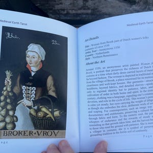 May include: An open book displaying a tarot card illustration from the Medieval Earth Tarot deck. The card features a portrait of a woman in Dutch folk costume from circa 1550, holding vegetables and a basket.