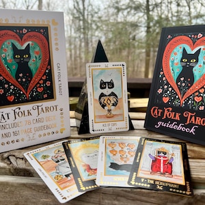 以下が含まれることがあります： Cat Folk Tarotセットには、金色の文字が書かれた白い箱、黒いガイドブック、猫のイラストが描かれたタロットカードが含まれています。箱にはCat Folk Tarot, includes 78 card deck and 161 page guidebookと書かれています。カードにはAce of Cups、Seven of Cups、The Hierophantが含まれています。