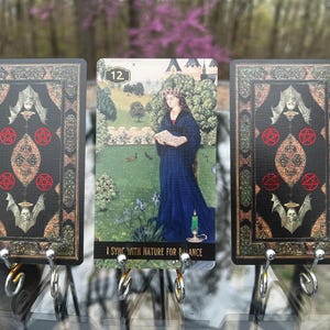 May include: Tarot card set featuring three cards with ornate black and gold borders. The central card depicts a woman in a blue gown in a garden, with the text "I SYNC WITH NATURE FOR BALANCE". The other cards have decorative symbols.