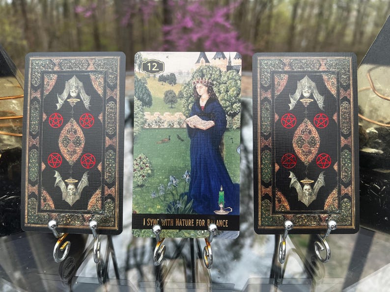 May include: Tarot card set featuring three cards with ornate black and gold borders. The central card depicts a woman in a blue gown in a garden, with the text "I SYNC WITH NATURE FOR BALANCE". The other cards have decorative symbols.
