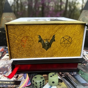 May include: A gold-colored box with a textured surface, featuring a bat skull emblem and pentagram symbols. The box sits atop a red-edged book with a red ribbon, surrounded by tarot cards and two green dice. The theme is occult and divination.