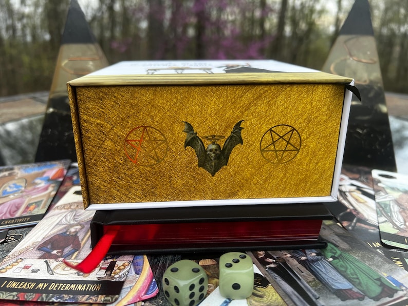 May include: A gold-colored box with a textured surface, featuring a bat skull emblem and pentagram symbols. The box sits atop a red-edged book with a red ribbon, surrounded by tarot cards and two green dice. The theme is occult and divination.