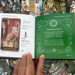 May include: An open book from the "Oracle of the Witches' Candle" deck, featuring a card with an illustration of a scene with figures and text. The opposite page has green text on a green background. The book is surrounded by other tarot cards.
