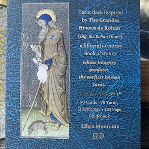 May include: A blue textured box for a tarot deck featuring a medieval illustration of a bearded figure with a staff and a small pig. The text reads: Tarot deck inspired by The Grandes Heures de Rohan, 90 Cards, 78 Tarot, 12 Astrology, 252 Page Guidebook, Libra Moon Inc.