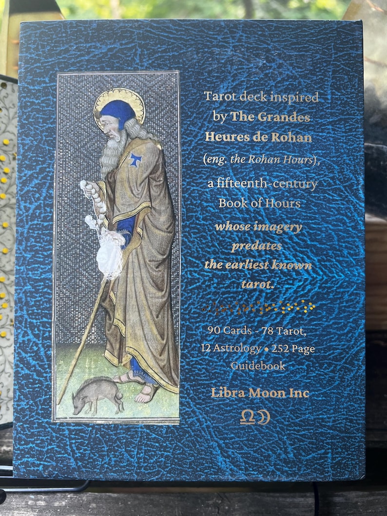 May include: A blue textured box for a tarot deck featuring a medieval illustration of a bearded figure with a staff and a small pig. The text reads: Tarot deck inspired by The Grandes Heures de Rohan, 90 Cards, 78 Tarot, 12 Astrology, 252 Page Guidebook, Libra Moon Inc.