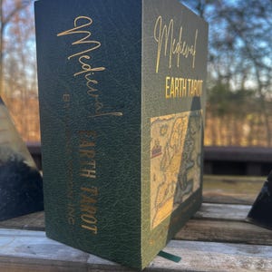 May include: A dark green tarot card box with gold lettering that reads "Medieval Earth Tarot." The box has a textured appearance and features a map design. The box is sitting on a wooden surface.