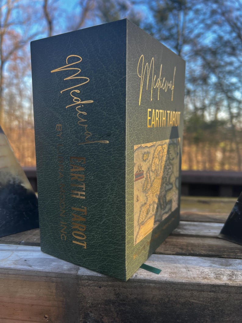 May include: A dark green tarot card box with gold lettering that reads "Medieval Earth Tarot." The box has a textured appearance and features a map design. The box is sitting on a wooden surface.