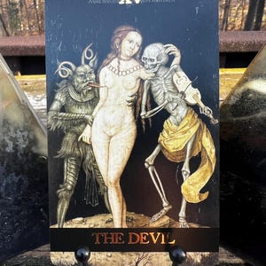 May include: A tarot card featuring a dark background with the text "THE DEVIL" at the bottom. The card depicts a nude woman, a skeleton playing a violin, and a devil-like creature. The top of the card has the Roman numeral XV.