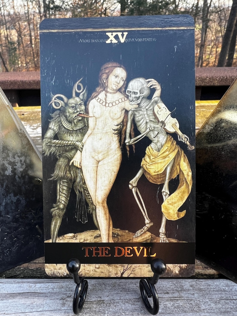 May include: A tarot card featuring a dark background with the text "THE DEVIL" at the bottom. The card depicts a nude woman, a skeleton playing a violin, and a devil-like creature. The top of the card has the Roman numeral XV.