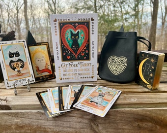 Könnte beinhalten: Ein Schwarz-Weiß-Katzen-Tarot-Deck mit einem Handbuch, einem schwarzen Beutel mit einem goldenen Herzdesign und einer einzelnen Karte, die eine schwarze Katze mit einer Mondsichel und Sternen zeigt. Die Karten haben goldene Akzente und das Handbuch hat eine schwarz-weiße Katze auf dem Cover.