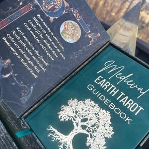 May include: A Medieval Earth Tarot Guidebook with a teal cover and gold lettering. The cover features a detailed white tree illustration. The open box reveals text and illustrations, including the phrases "Roots entwine in sacred stone" and "Medieval Earth".