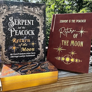 Può includere: Due libri e un mazzo di carte con bordi dorati. I libri sono intitolati "Serpent and the Peacock: Return of the Moon". Uno è nero con dettagli argentati ornati, l'altro bordeaux con lettere dorate. Il mazzo di carte ha bordi dorati.