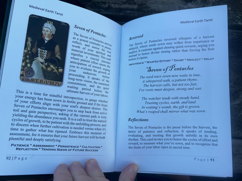 May include: Open book with a Medieval Earth Tarot spread. The left page features the "Seven of Pentacles" card with an illustration of a person and text. The right page has text about the card's reversed meaning.
