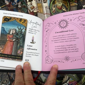May include: An open book displaying a tarot card reading. The left page features a card with an illustration of two figures embracing and a horned creature. The right page has text about unconditional love, with a pink background.