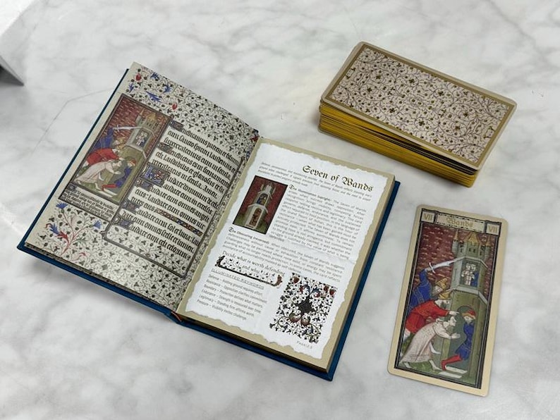 May include: An open book with medieval-style illustrations and text, a deck of tarot cards with gold edges, and a single Seven of Wands tarot card. The book pages feature intricate floral borders, Latin text, and a detailed description of the card.